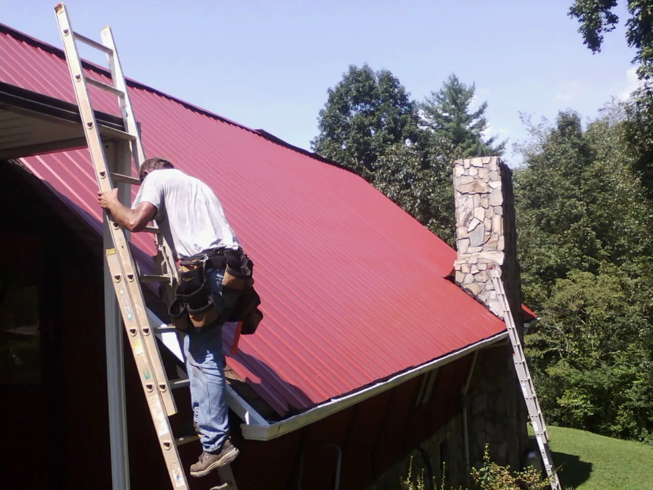Roofer on a ladder beside a red metal roof and stone chimney — Soffit & Fascia Repair by Roofing Brothers in Sekiu
